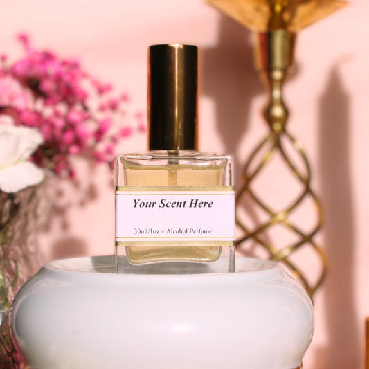 A square clear glass perfume bottle with a golden cap, labeled 'Your Scent Here - 30ml/1oz - Alcohol Perfume'. The liquid inside has a slight golden hue. Set against a pastel pink background, the product is complemented by soft focus elements such as pink flowers and a gold candlestick, reflecting a delicate and sophisticated ambiance.