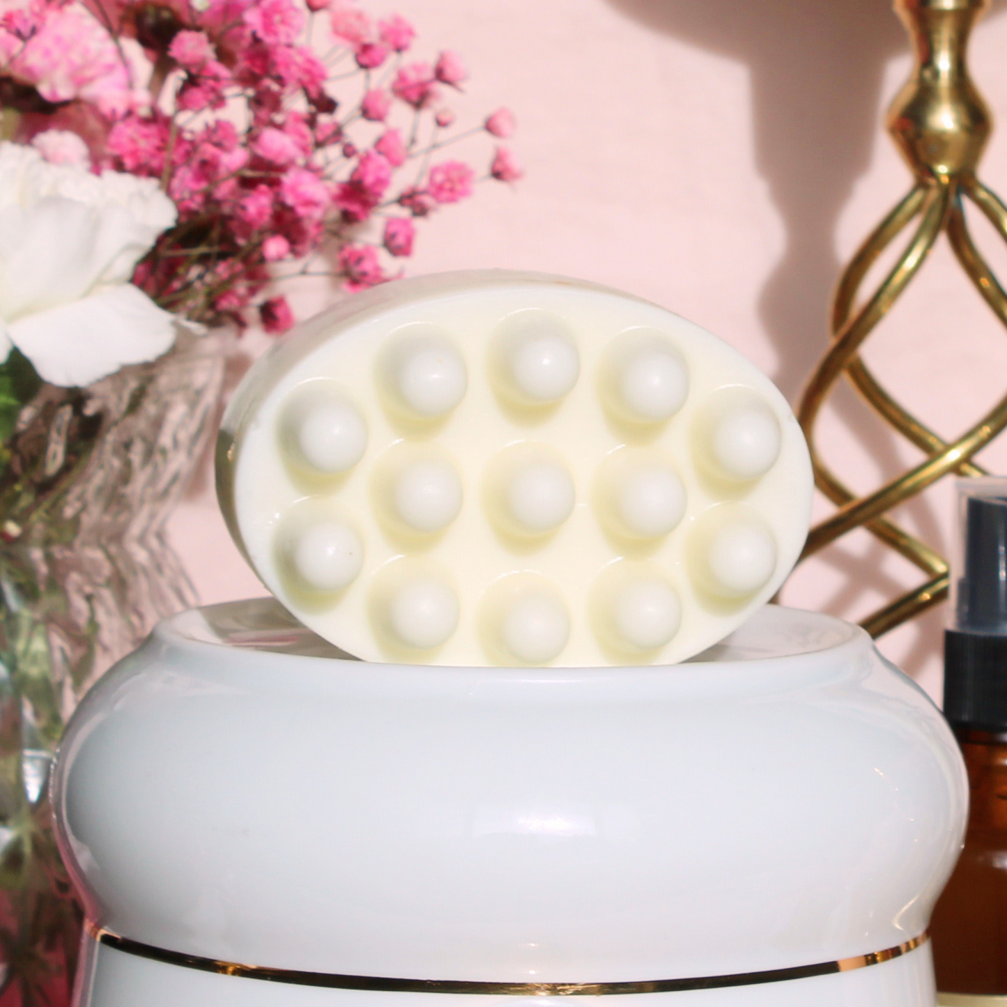 A circular white massage soap bar with multiple raised bumps on its surface, resting on top of a white ceramic jar. The jar is placed on a table with a pink backdrop, next to a bouquet of pink flowers and a blurred amber glass bottle.