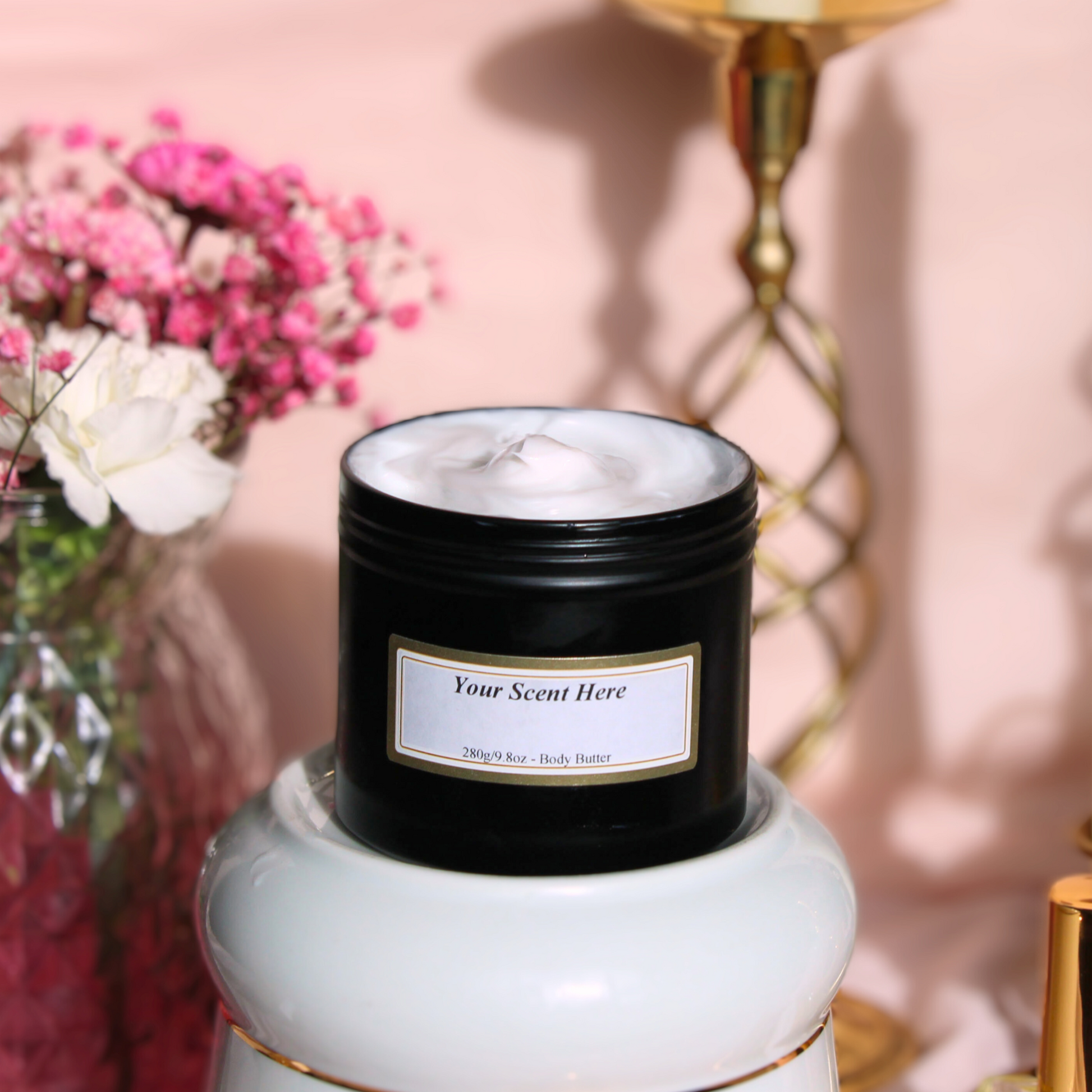 An open black jar of body butter, labeled 'Your Scent Here - 280g/8oz - Body Butter' with a smooth, creamy texture visible at the top. The jar rests on a light-colored surface with a soft pink wall behind it, accompanied by a bouquet of pink flowers and a golden candle holder, adding to the product's luxurious and soothing aesthetic.