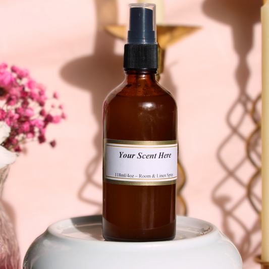 A 4oz (118ml) amber glass spray bottle labeled 'Your Scent Here' for room and linen, displayed on a white stand with a backdrop of soft pink and a decorative gold element in the background. A small bouquet of pink flowers is partially visible to the left.