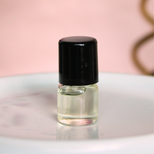 A small clear glass bottle with a black cap, containing a pale yellow liquid, which is oil perfume, displayed on a white surface with a soft pink background.