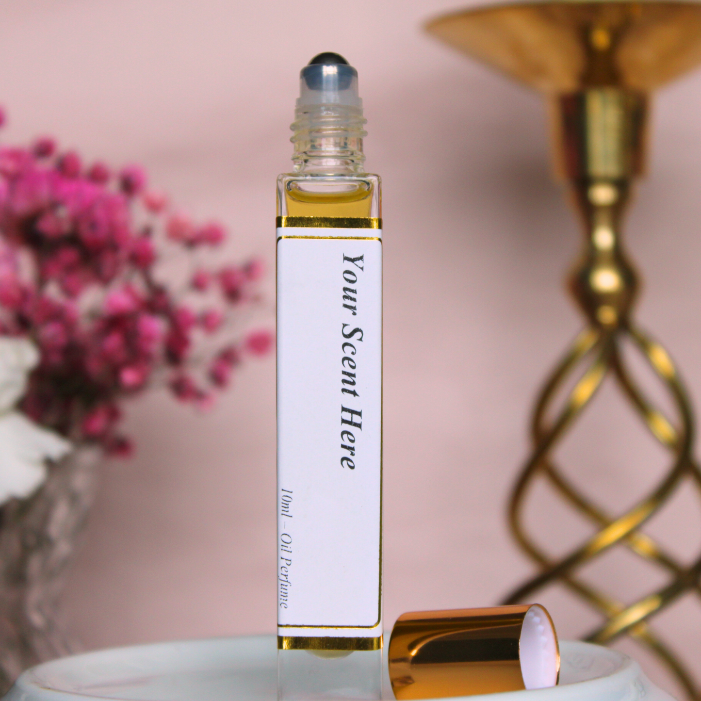 Close-up of an open clear glass roller bottle with a gold-colored cap beside it, showing a rolling ball applicator. It's labeled 'Your Scent Here' and contains a golden-yellow liquid, likely oil perfume. The background features soft pink tones with a cluster of pink flowers and a part of a gold candlestick, contributing to the elegant presentation of the product.