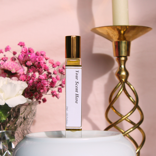 A tall, slender clear glass roller bottle with golden cap, labeled 'Your Scent Here', filled with yellow liquid, an oil-based perfume. The bottle is perched on a white surface with a bouquet of pink flowers and a white flower to the left, and a golden candlestick with a white candle in the background, all against a pale pink backdrop.