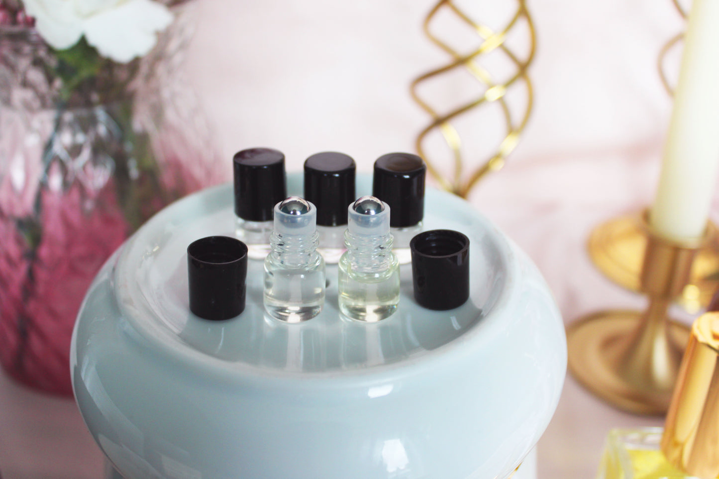 An array of small clear glass bottles with black caps, each filled with a clear to pale yellow liquid, displayed on a white ceramic dish. The dish is set against an elegant background with pink hues, a vase with white flowers, and a golden decorative element, creating an ambiance of luxury and tranquility for these sample-sized oil perfumes